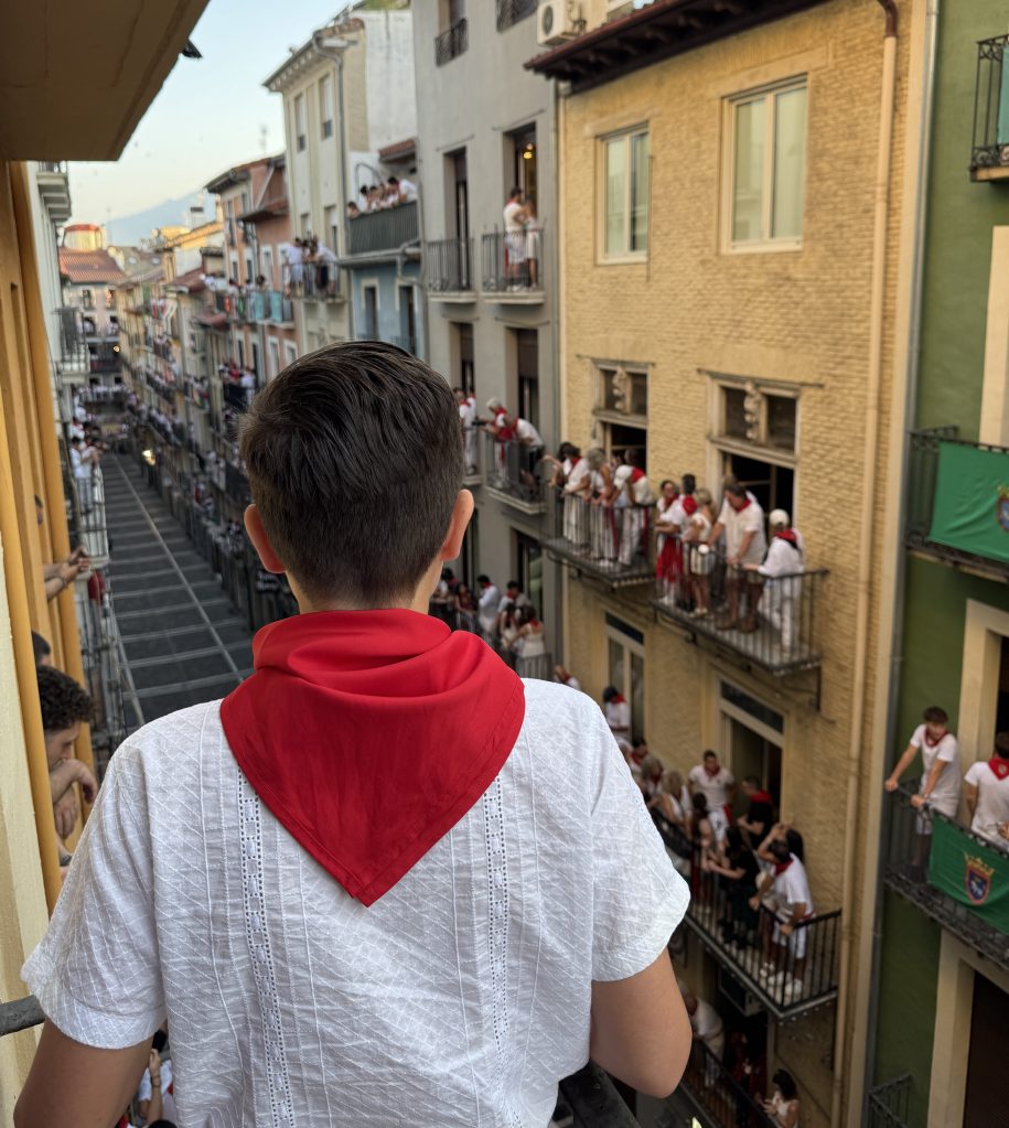 A young person wearing a white shirt and a red scarf stands on a balcony overlooking a bustling street filled with people celebrating, some on balconies wearing white clothing, during a vibrant festival.