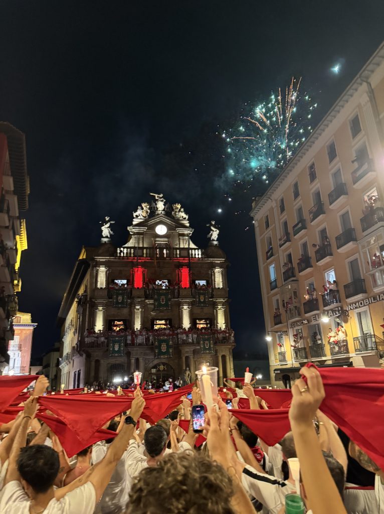 Crowd celebrating at night with red flags and candles in front of a historic building, fireworks in the sky above.