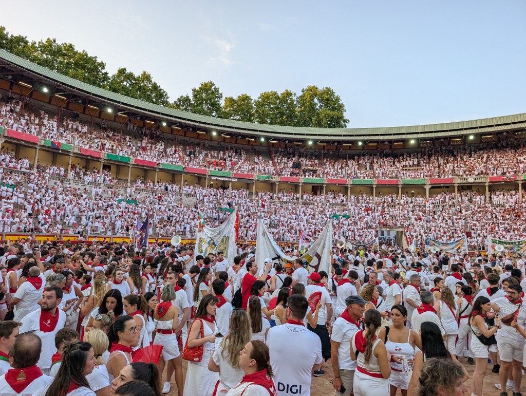A large crowd gathered in an arena, dressed in white with red accents, celebrating a festive event. Banners and flags are displayed, creating a vibrant atmosphere.