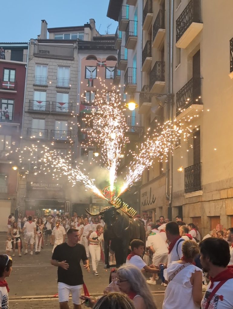 A lively street scene during a festival, featuring people in white clothing with red accents celebrating around a traditional figure resembling a bull, which is emitting sparks and fireworks.